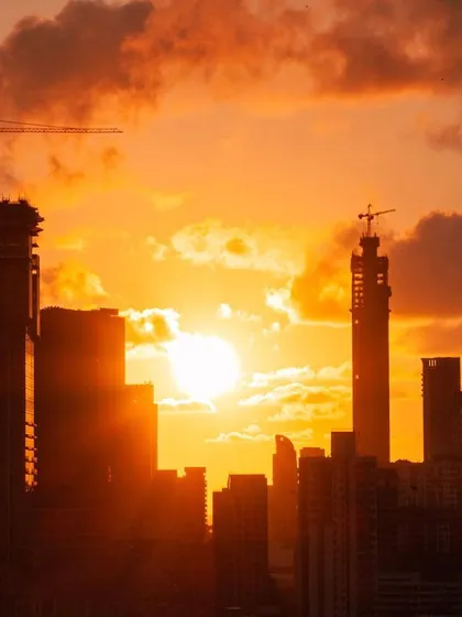 The sun sets between the silhouettes of Mumbai's buildings, with cranes visible on under-construction towers, all under a fiery orange sky.