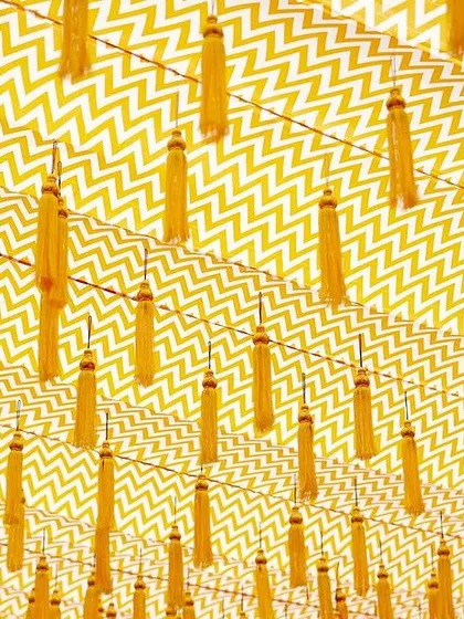 A creative ceiling treatment using a chevron-patterned fabric with hanging yellow tassels, adding a modern graphic element to the traditional Haldi decor.