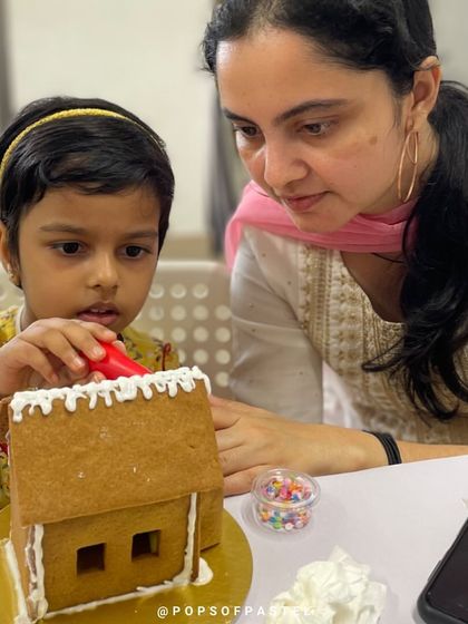 A mother helps her daughter carefully apply icing to the walls of her gingerbread house. It's a perfect parent-child bonding activity.