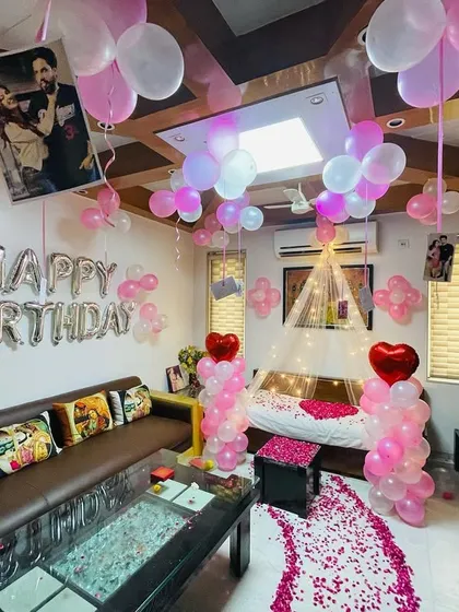 A full room decoration for a birthday surprise. The setup includes balloons on the ceiling, a "Happy Birthday" foil banner, and a path of rose petals leading to the decorated bed.