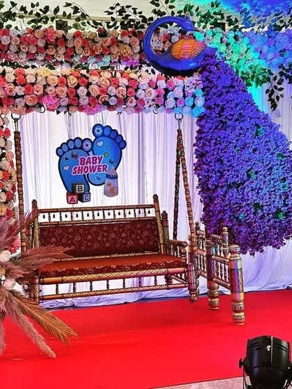 A vibrant peacock-themed decoration for a Dohale Jevan. The setup includes a floral swing, a large peacock figure made from purple flowers, and gender-themed cutouts with pink and blue balloons.