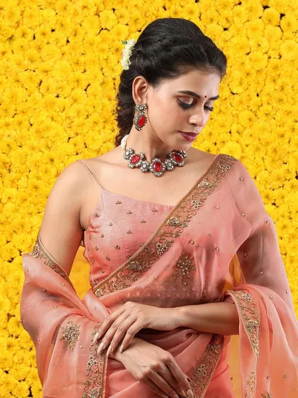 A close-up portrait of the model, showcasing the beautiful peach saree and the statement red stone necklace.