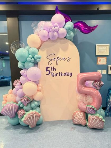 A mermaid-themed 5th birthday with a custom backdrop and a large number "5" foil balloon. The balloon garland features shades of teal, lavender, and peach with seashell accents.