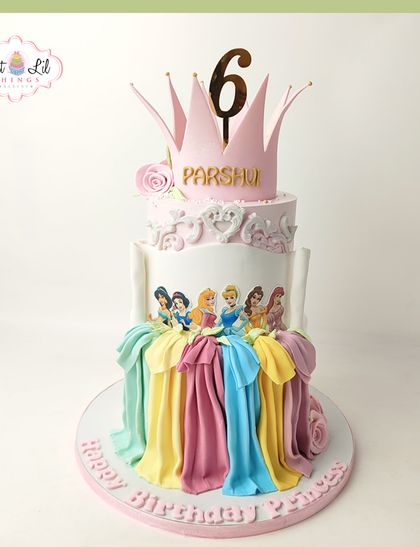 A royal celebration for a Disney Princess fan. This grand three-tiered cake is decorated with the flowing gowns of multiple princesses and topped with a beautiful pink crown for a 6th birthday.