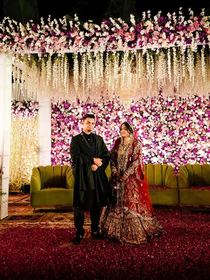 The couple stands proudly on their beautifully decorated stage. The combination of the floral wall, hanging wisteria, and plush green sofas creates a look of pure elegance.