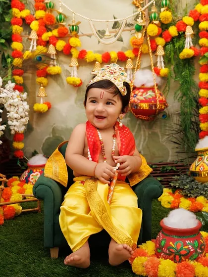 A happy baby Krishna sits on a miniature chair, surrounded by the vibrant colors of the Janmashtami festival.