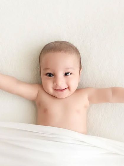 A happy baby lies on a white blanket, stretching his arms out wide.