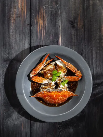 A dramatic overhead shot of a crab dish in a dark sauce, using shadows to create a high-end, artistic feel for the Zarin menu.