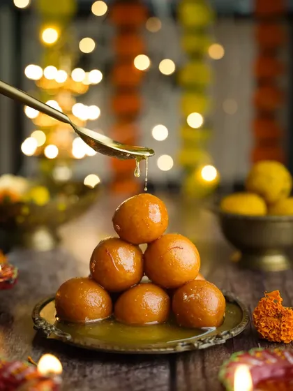 Happy Diwali. A classic festive shot of gulab jamuns stacked in a pyramid, with a spoon pouring syrup over them. The diyas and flower garlands in the background complete the scene.
