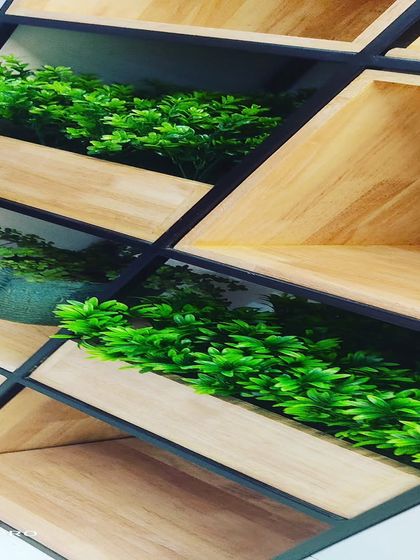A close-up of a geometric shelving unit with small artificial plants. The plants are placed inside the angled wooden boxes, adding a touch of green to the modern design.