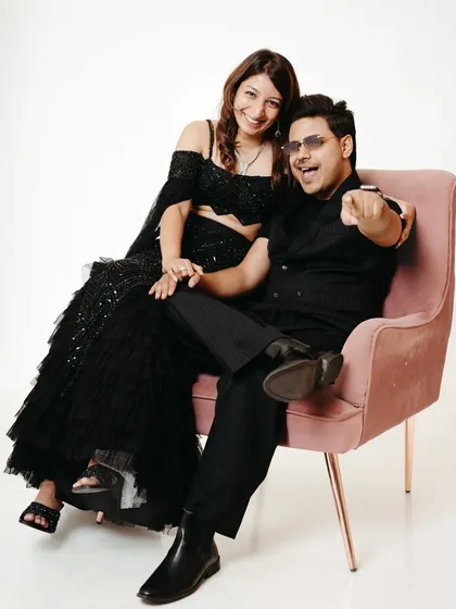 This studio portrait has a cool, editorial feel. The couple sits together on a pink chair, with the man pointing confidently at the camera, capturing a moment of shared fun and style against a simple white background.