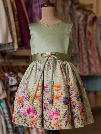 This sage green dress features a beautiful border print of wildflowers. The simple satin bodice and delicate bow at the waist let the stunning floral skirt take center stage.