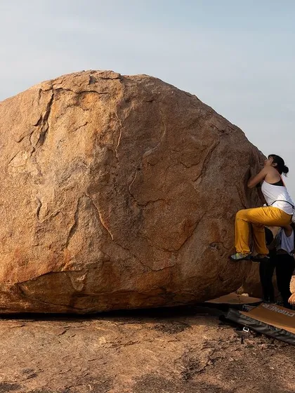 A wide shot showing a bouldering session in full swing. The climber on the rock is the focus, but the community on the ground is her foundation.