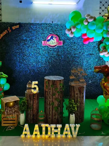 A Jurassic Park inspired dinosaur theme with a dark grass wall and wood-textured pedestals. Foil dinosaur balloons and marquee letters add to the prehistoric vibe.