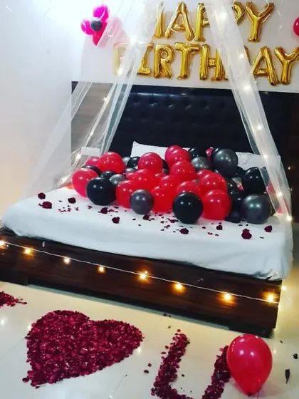 A romantic birthday surprise for a wife. The room features a canopy with fairy lights, red and black balloons, and an "I Love You" message made from rose petals.