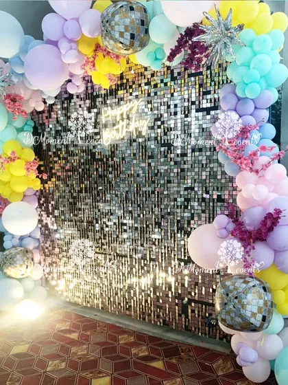 A dazzling photo backdrop featuring a silver sequin wall framed by a pastel rainbow balloon arch. Disco balls and starburst accents add a touch of sparkle and fun to the rainbow theme.