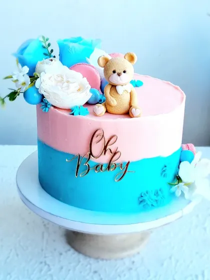 A "He or She" baby shower cake with a pink and blue design. The cake is topped with a cute fondant teddy bear, macarons, and sugar flowers.