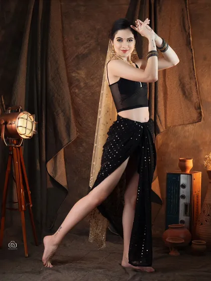 An Indo-western look combining a modern black top and draped skirt with a traditional sheer dupatta. The studio set, complete with rustic props, adds a unique, theatrical feel to the shot.