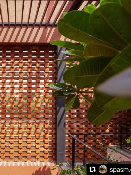 A close-up view of the brick jali wall with the leaves of a frangipani tree in the foreground. The contrast between the rigid geometry of the brick and the organic form of the leaves is a key part of the design.