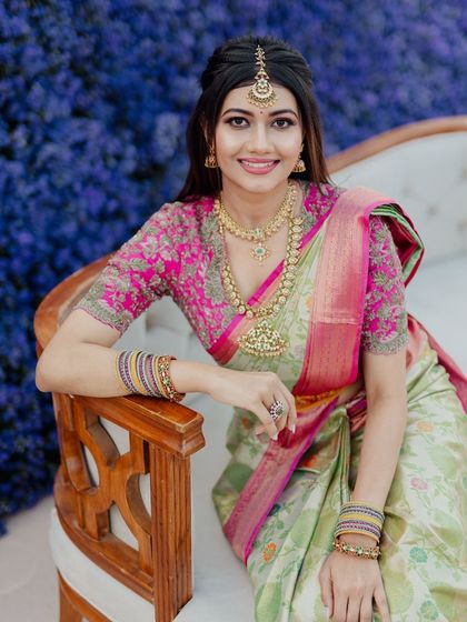A beautiful bride seated against a backdrop of purple flowers. Her makeup is soft and romantic, with a pink blouse adding a pop of color.