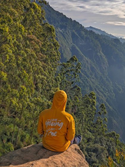 A traveler in our "Live Young, Live Free" hoodie sitting at Dolphin's Nose viewpoint in Kodaikanal, enjoying the spectacular valley view.