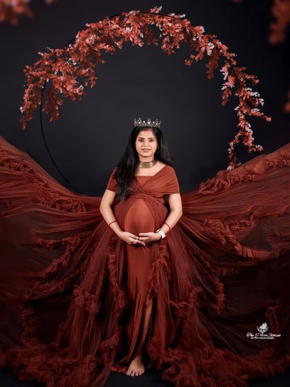 A powerful and regal solo portrait. The mom-to-be wears a crown and a dramatic brown gown, framed by a beautiful floral arch against a black background.