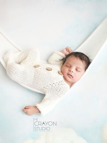 A creative composite image showing a newborn sleeping peacefully in a soft white hammock against a sky-blue backdrop with fluffy clouds.