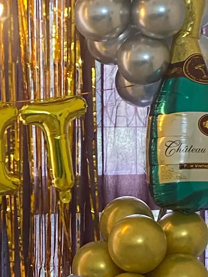 Sometimes, a few key elements are all you need. This close-up shows a large champagne bottle foil balloon paired with gold letter balloons against a shimmering gold fringe curtain, perfect for an elegant anniversary or success party.