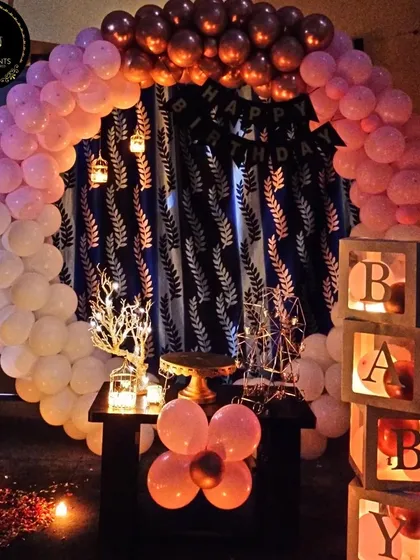 A cozy and warm birthday setup ideal for a small room or corner space. The pink, white, and rose gold balloon arch frames a simple backdrop, complemented by candle-lit 'BABY' blocks for a soft, intimate glow.