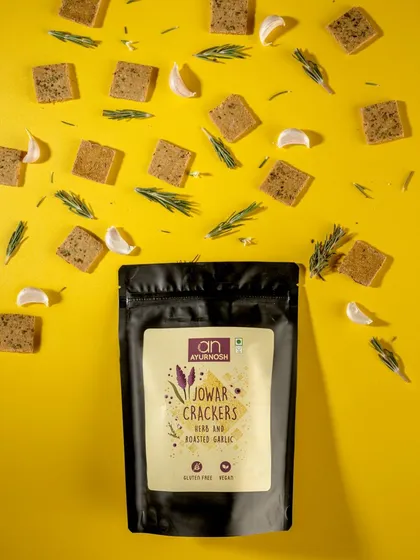 A vibrant flatlay for Ayurnosh's Jowar Crackers. The "explosion" of crackers, garlic, and rosemary on a bright yellow background is energetic and effectively communicates the "Herb and Roasted Garlic" flavor.
