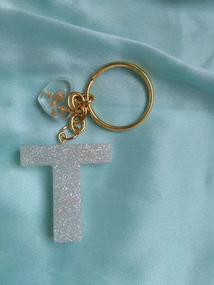 A simple and elegant initial keychain. This 'T' is made with a sparkling white glitter resin and comes with a small, clear heart charm filled with gold flakes, attached to a gold-plated ring.