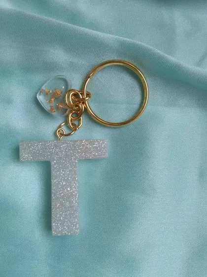 A simple and elegant initial keychain. This 'T' is made with a sparkling white glitter resin and comes with a small, clear heart charm filled with gold flakes, attached to a gold-plated ring.