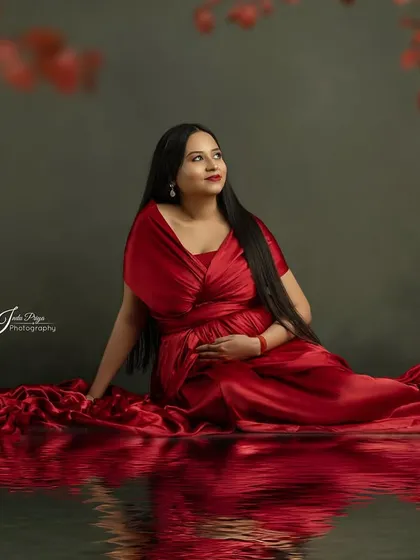 A beautiful composition featuring a mom-to-be in a red gown, seated with a water reflection effect. The soft red leaves at the top of the frame add depth and a touch of nature.