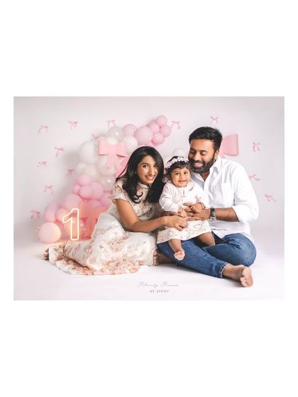 A smiling family celebrating their daughter's first birthday. The pink and white balloon decor, along with a glowing "1" sign, creates a perfect party atmosphere in the studio.