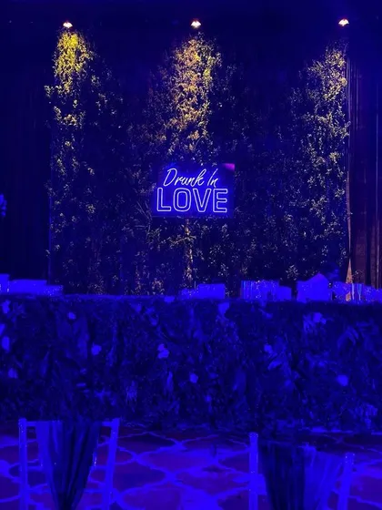 A stylish bar setup for a sangeet night. A neon sign saying "Drunk in Love" is set against a green foliage wall, illuminated by cool blue lighting to match the party's vibe.