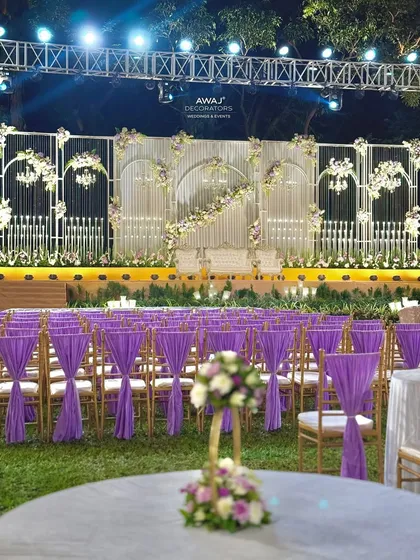 This dreamy outdoor wedding stage combines soft lavender and white florals with elegant white structures, perfectly illuminated against the night sky for a sophisticated and romantic sangeet or reception.