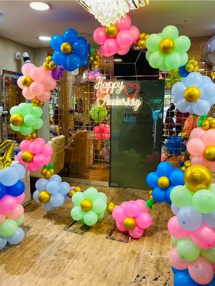 A fun and colorful flower-themed anniversary decoration. The balloon arch is made of balloon daisies in various colors, with a "Happy Anniversary" neon sign.
