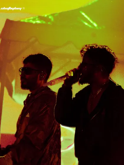 A silhouette of Seedhe Maut against a hazy yellow and green background. This artistic shot captures the duo's profile and the smoky atmosphere of the venue.