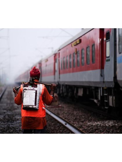 A railway worker in a bright orange vest walks along the tracks next to a train. The image captures the essential, often unseen, labor that keeps the country moving.