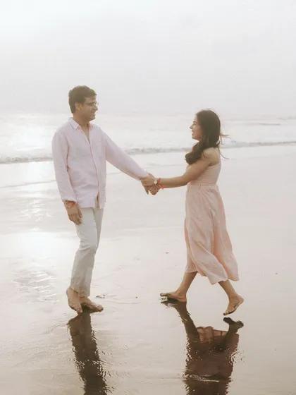 Ritu and Tarush holding hands as they walk along the shoreline. The soft sunset light and their reflections in the wet sand create a dreamy, romantic atmosphere for this pre-wedding photograph.