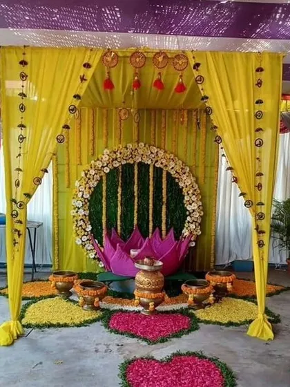 This unique Haldi decor features a central lotus-shaped seating area. The backdrop is a floral ring on a green wall, surrounded by a canopy of yellow drapes and flower arrangements.