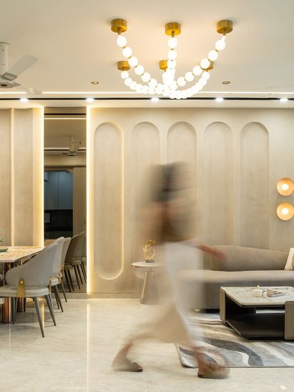This luxurious living-dining space embraces fluidity through its curved wall paneling and layered lighting. A sculptural chandelier anchors the ceiling, while soft neutral tones and brushed gold accents create a seamless blend of modern sophistication.