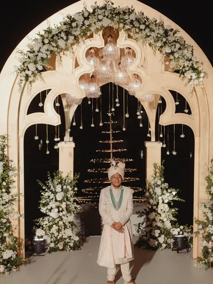 The groom awaits his bride, standing under the magnificent floral arch on the over-water Varmaala stage. The structure's intricate design and the soft glow of the lights create a perfect picture of anticipation and elegance.