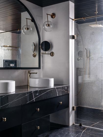 The master bathroom in our Ebony & Ivory project is a moody masterpiece. It features a black marble vanity, twin vessel sinks, an arched mirror, and modern brass fixtures, all set against a dark, wood-paneled ceiling.
