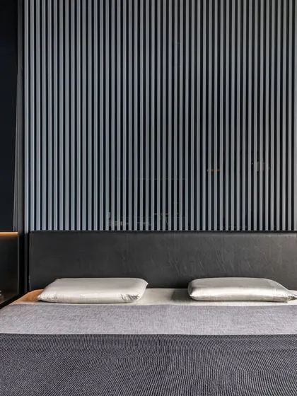 This bedroom showcases our focus on integrated design elements. A custom light installation in Nero metal unfolds from the wall to become a functional bedside table. The headboard wall, crafted from grey ribbed wooden slats against a smoked glass background, creates an illusion of depth and breathability through subtle reflections.