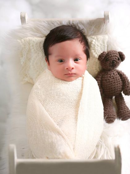 A wide-eyed newborn lies in a miniature bed, accompanied by a tiny teddy bear. The dreamy, cloud-like setting adds a touch of whimsy to this sweet shot.