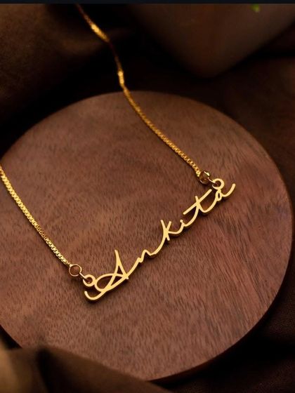 The 'Ankita' script necklace, presented on a wooden base, highlights the warm glow of the gold finish. Each nameplate is carefully crafted to be a personal treasure.