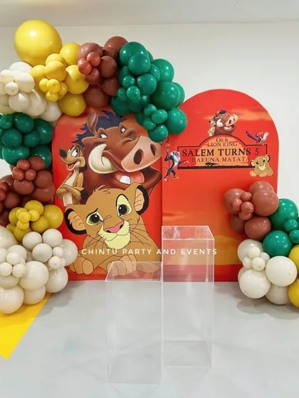 A Lion King themed 5th birthday party decoration. The setup uses earthy tones of green, brown, and yellow balloons to frame a backdrop featuring Simba and friends.