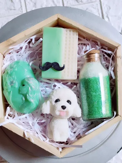 A unique gift hamper featuring a dog-shaped soap, a lady-face soap, a mustache soap, and green apple bath salts in a hexagonal box.