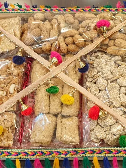 A square tray hamper for Lohri, filled with an assortment of traditional winter sweets like gajak and chikki, decorated with colorful pom-poms.